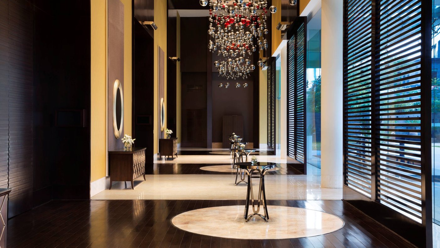Pre-function corridor at JW Marriott Bengaluru with dark timber louvred screens, a chandelier of clustered silver and red spheres, and cocktail tables on inset stone circles
