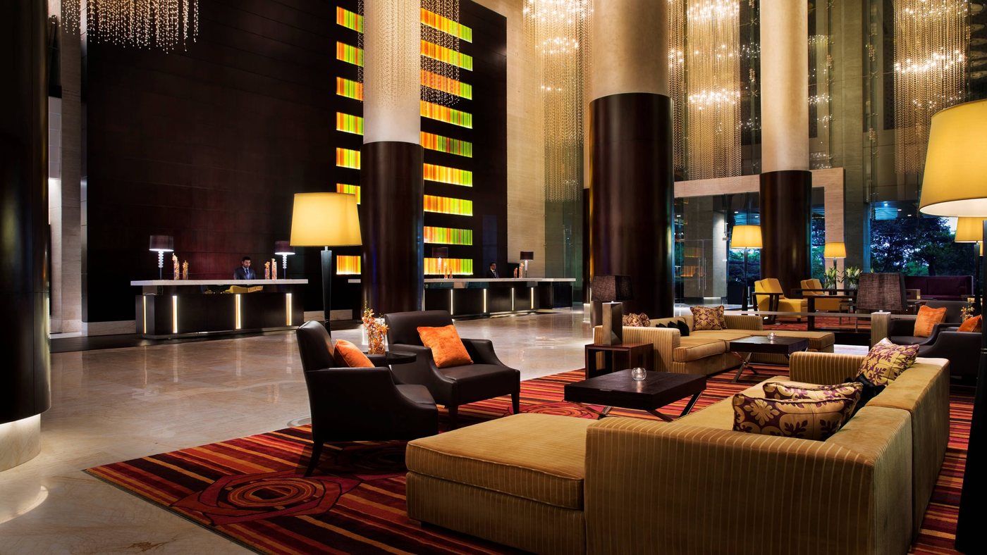 The lobby at JW Marriott Bengaluru — double-height columns with cascading crystal-strand chandeliers, a backlit coloured glass panel behind reception, and low seating on a striped red carpet