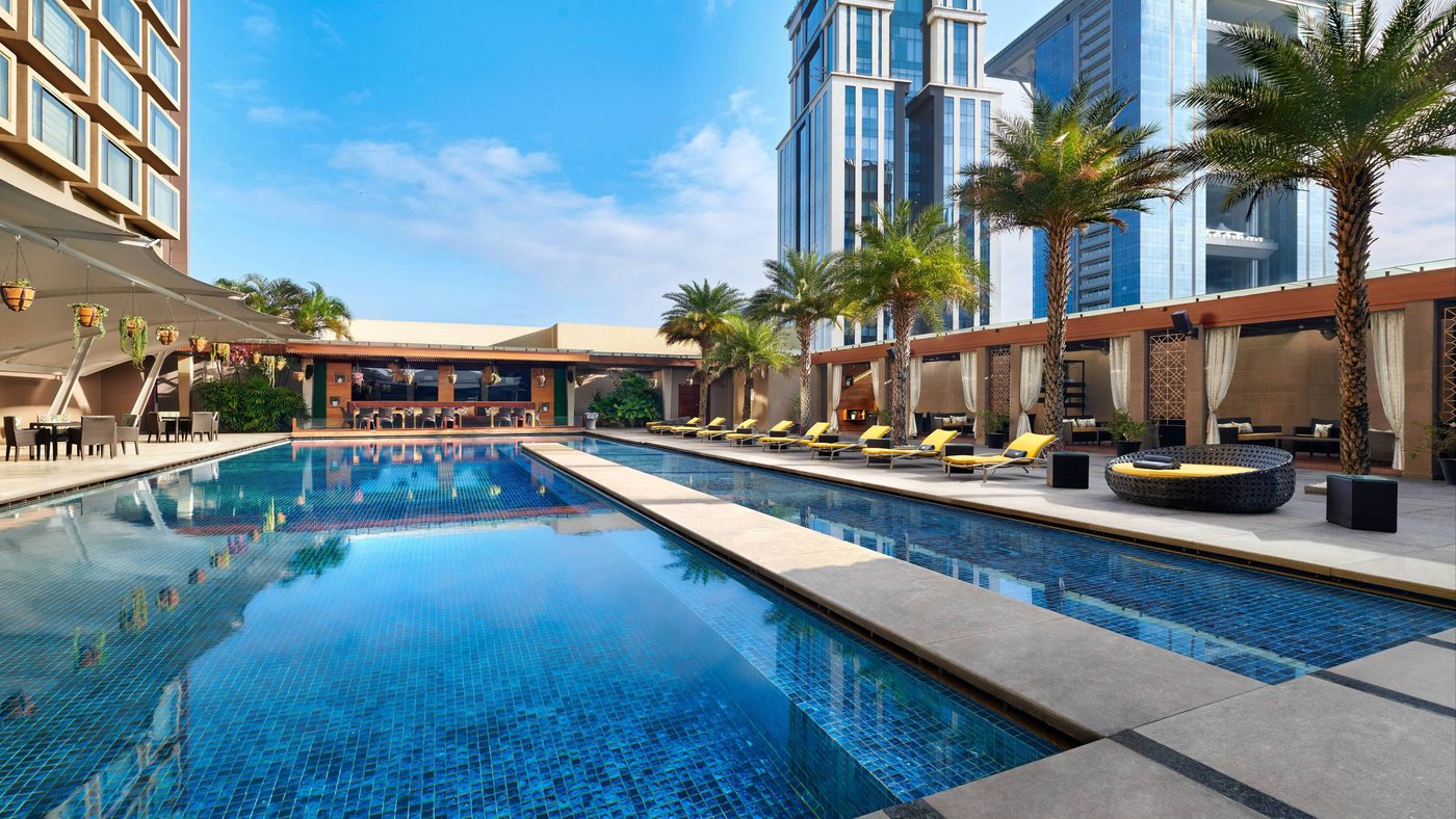 The pool deck at JW Marriott Bengaluru — twin mosaic-tiled pools with sun loungers, date palms and cabanas, with the Vittal Mallya Road office towers behind
