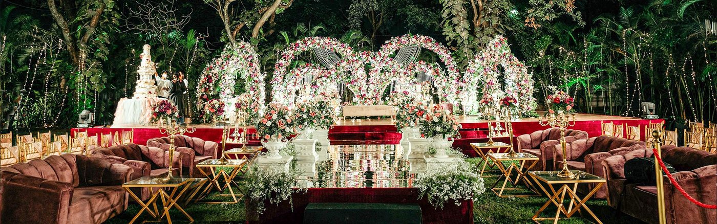 Outdoor wedding stage at The LaLiT Ashok Bangalore with a white and gold floral mandap and low seating, set against dense bamboo and garden planting