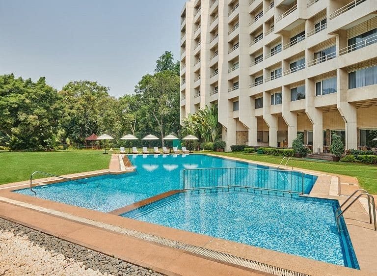 The outdoor pool at The LaLiT Ashok Bangalore with a lawn and mature trees on one side and the hotel's stepped facade rising behind