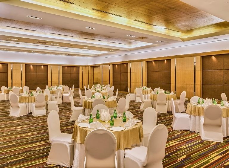 Banquet hall at The LaLiT Ashok Bangalore set with gold-linen round tables under a coffered gold ceiling and timber wall panelling