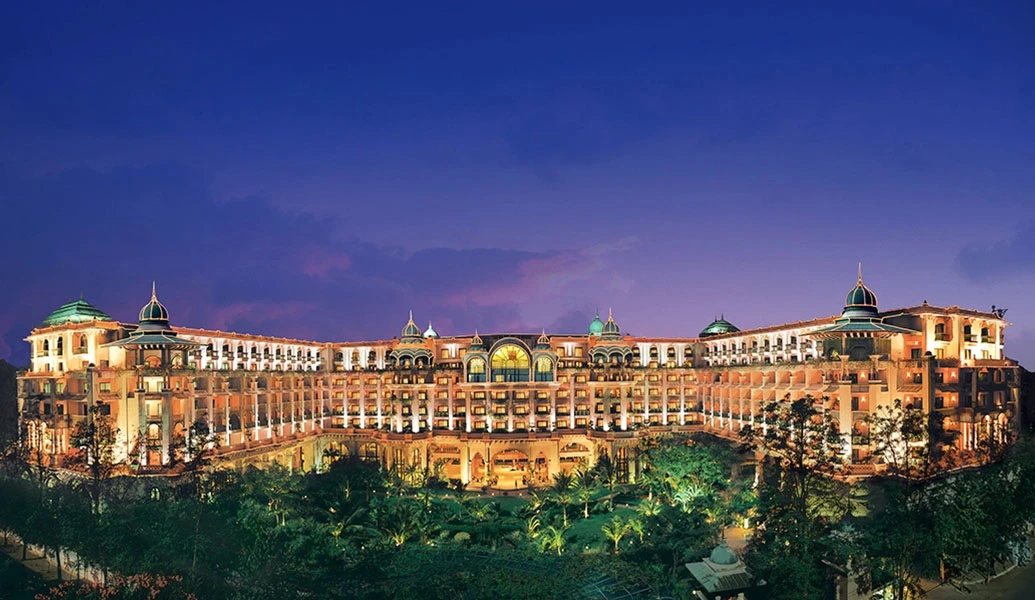 The Leela Palace Bengaluru floodlit at dusk against a violet sky — the full symmetrical pink facade with green domes and the central arched glass atrium, the porte-cochère lit below and the gardens dark around it