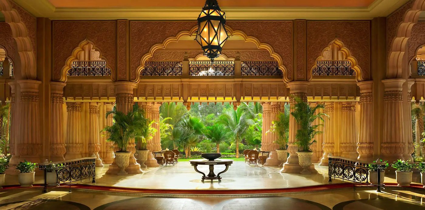 The pillared entrance hall of The Leela Palace Bengaluru looking out to the gardens — carved sandstone columns and a scalloped arch with fretwork balustrades above, a single lantern hanging over a stone centre table, and palms and lawn framed in the opening beyond