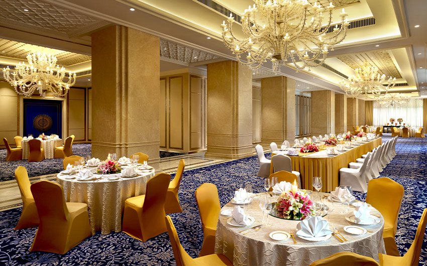 The Maharaja Ballroom at The Leela Palace Bengaluru set for a banquet — marble-clad columns down the centre, crystal chandeliers on a coffered gold ceiling, blue floral-patterned carpet, and gold-covered round tables with white place settings beside a long buffet run