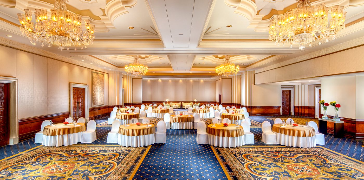 The Grand Ballroom at The Leela Palace Bengaluru set with gold-topped round tables and white chair covers — a pillarless room with a sculpted cream ceiling, four crystal chandeliers, timber dado panelling and two large blue-and-gold patterned rugs