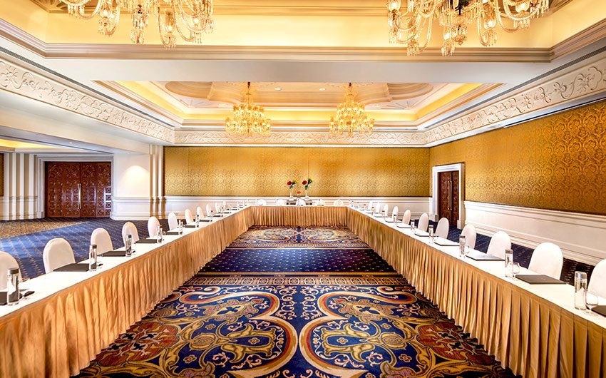 The Royal Ballroom at The Leela Palace Bengaluru laid out in a hollow-square U with gold skirted tables and white chairs, under crystal chandeliers and a carved white cornice, with gold damask walls and carved timber doors