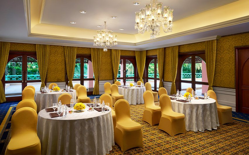Diya at The Leela Palace Bengaluru set for a small banquet — round tables with yellow chair covers under two crystal chandeliers, gold damask walls, and a run of arched French doors with gold curtains opening onto greenery and a turquoise balustrade