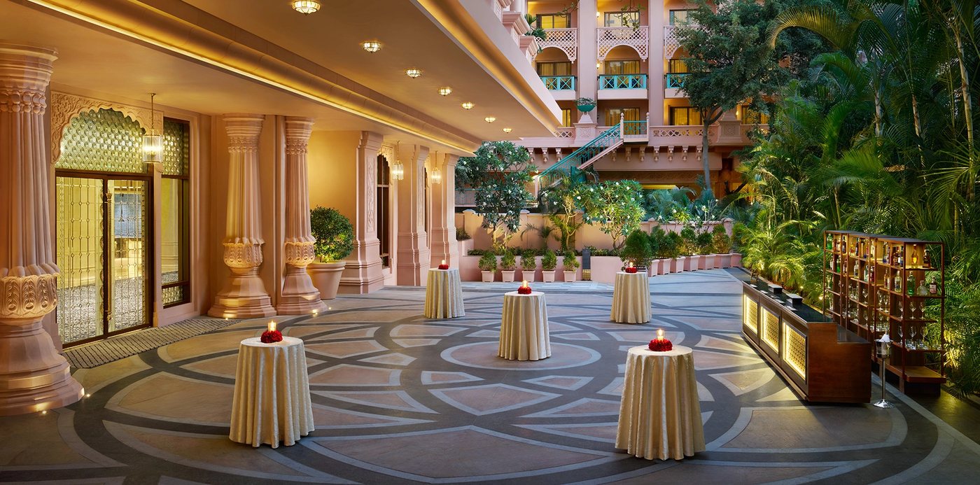 The Lotus Courtyard at The Leela Palace Bengaluru dressed for an evening cocktail reception — carved sandstone columns and a jaali screen along the covered edge, a stone medallion floor set with candlelit high tables, a lit bar on the right and the pink palace wings with planted balconies rising behind
