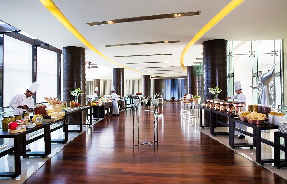 A live-station buffet set up in a corridor at Bengaluru Marriott Whitefield — chefs at long counters along a polished wooden floor beneath a sculpted ceiling