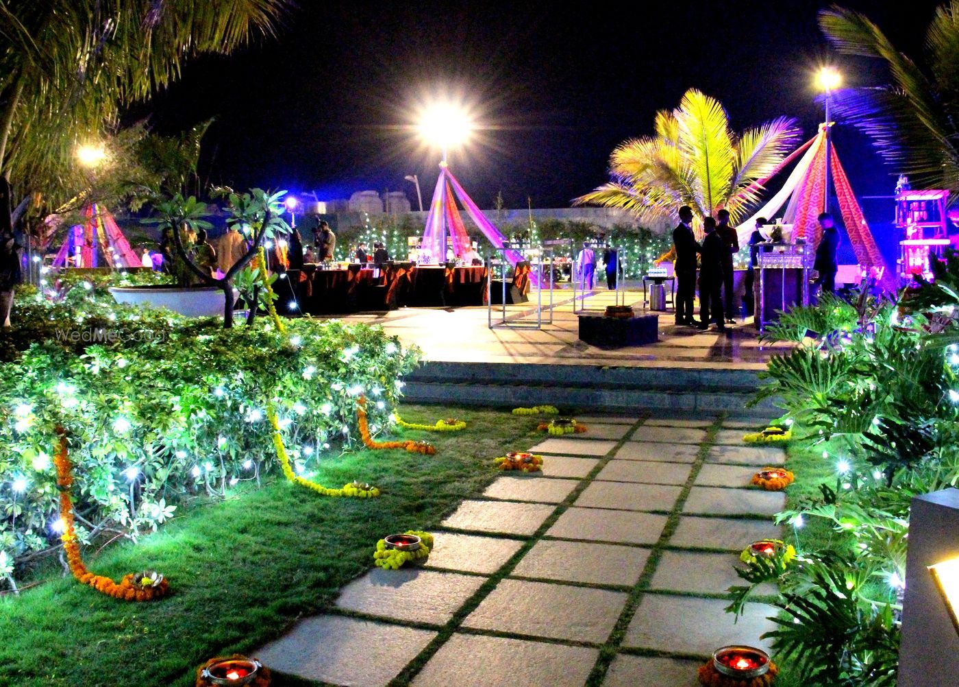 The pool deck at Bengaluru Marriott Whitefield dressed for an evening function — lit canopies and a raised platform beyond a paved path lined with diyas and marigold, palms uplit around it