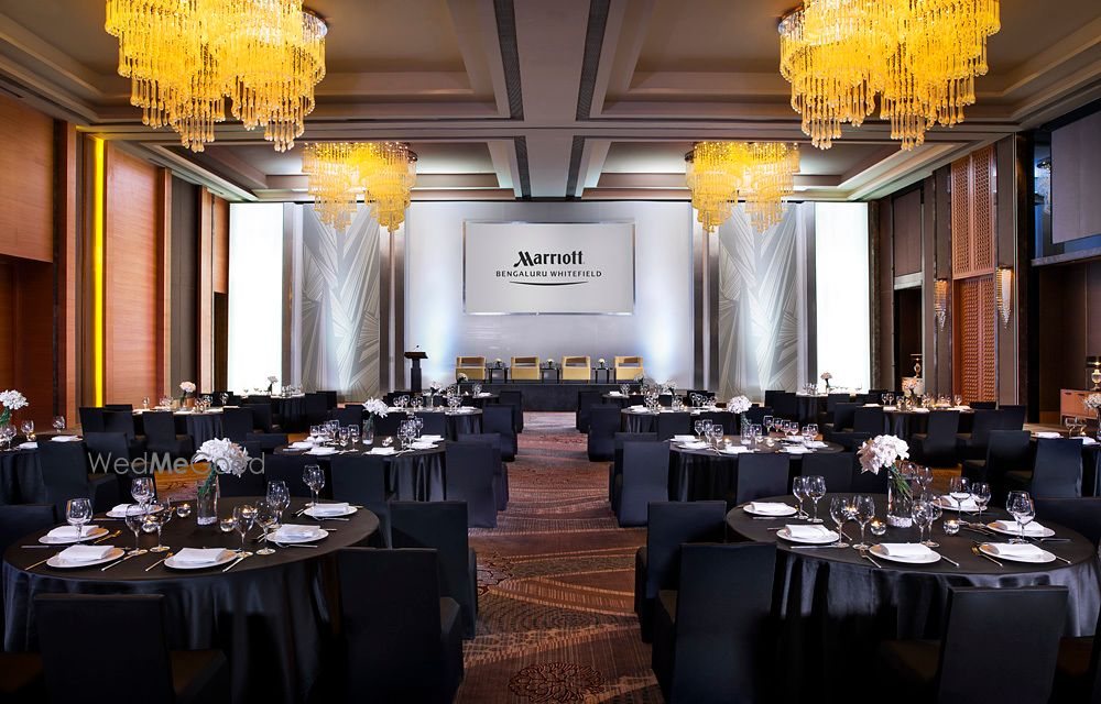 The Grand Ballroom at Bengaluru Marriott Whitefield set for a seated dinner — round tables in black linen under gold tiered chandeliers, with the hotel's LED wall lit at the front