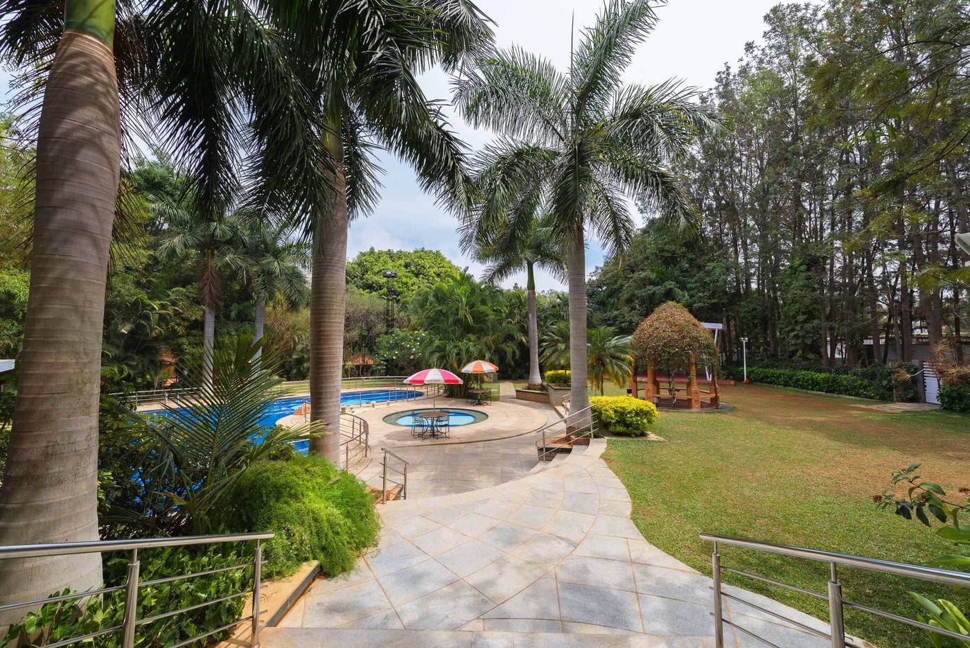 The swimming pool at MG Magnus seen from a stepped stone path — blue pool and circular plunge pool with parasols, a thatched gazebo, royal palms and open lawn beyond
