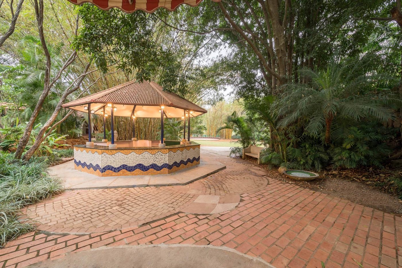The octagonal garden bar at MG Magnus — a mosaic-tiled counter under a conical tiled roof with lantern lighting, on brick and stone paving among trees
