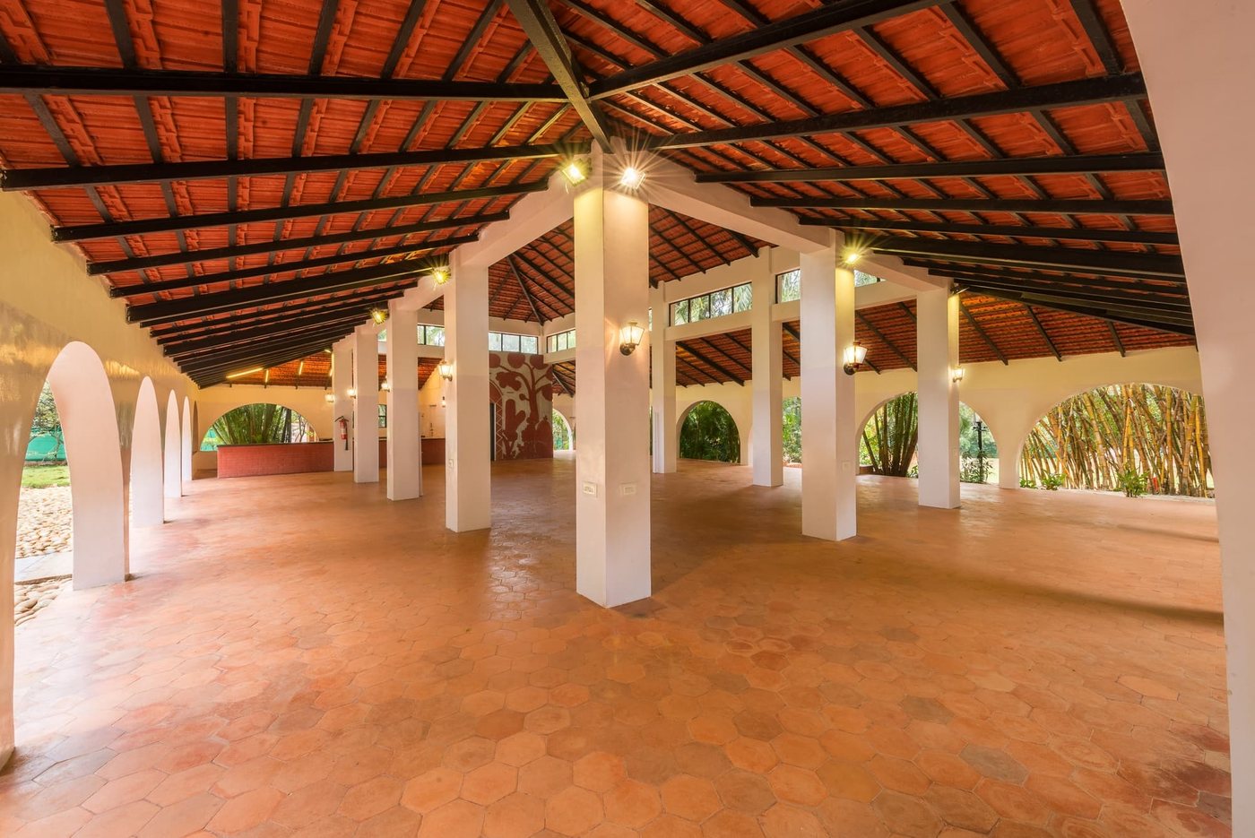 The open-sided dining pavilion at MG Magnus — white plastered columns and arches under a Mangalore-tiled roof on dark timber rafters, terracotta hexagonal tile floor, bamboo grove visible through the arches
