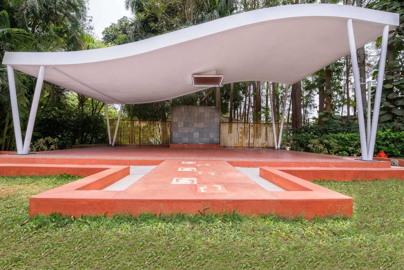 Performance stage at MG Magnus: a terracotta-red plinth under a curved white tensile canopy on splayed steel props, with a grey stone backdrop wall, set on the lawn