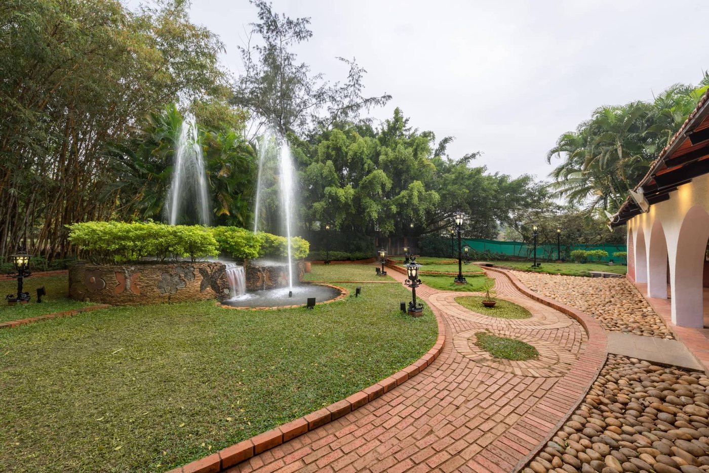 Fountain garden at MG Magnus with a raised stone basin, brick pathways, cast-iron lamp posts, bamboo and palms, and the arched pavilion to one side