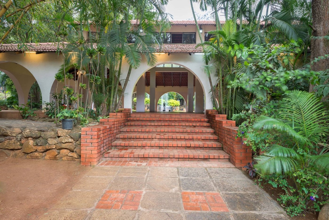Brick steps rising to the arched pavilion at MG Magnus, framed by areca palms and bamboo, with the garden fountain visible through the arches