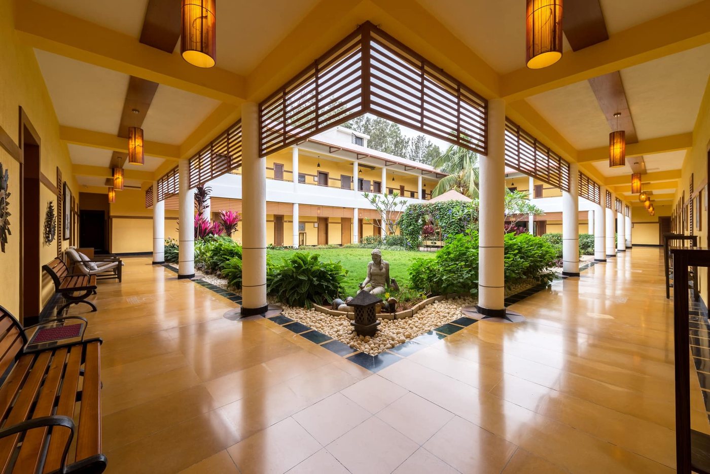 The Lotus accommodation block at MG Magnus — a double-height yellow-walled corridor with white columns and timber louvres running around a planted courtyard with a seated stone sculpture