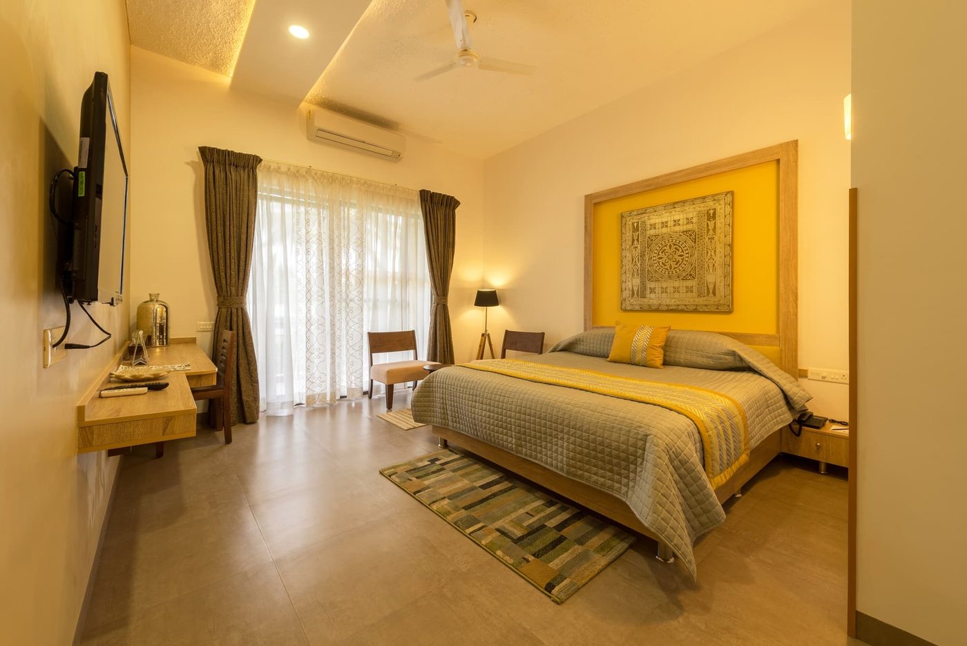 A guest room at MG Magnus with a yellow headboard panel framing tribal artwork, sheer curtains onto the balcony, air-conditioning and a desk