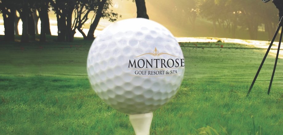 Montrose-branded golf ball on a tee on the resort's fairway, the tree line of the estate behind in low sun