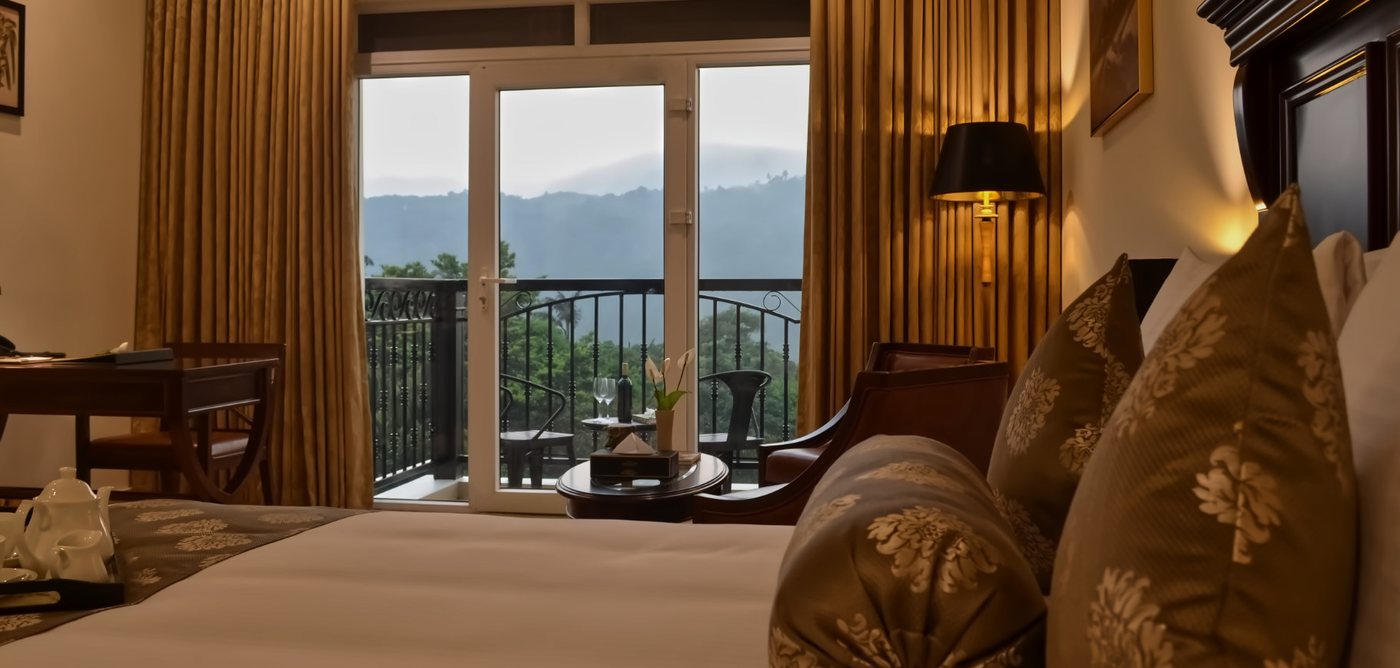 Suite at Montrose Golf Resort with French doors open to a balcony and the Western Ghats ridgeline beyond, dark timber bed and gold drapes