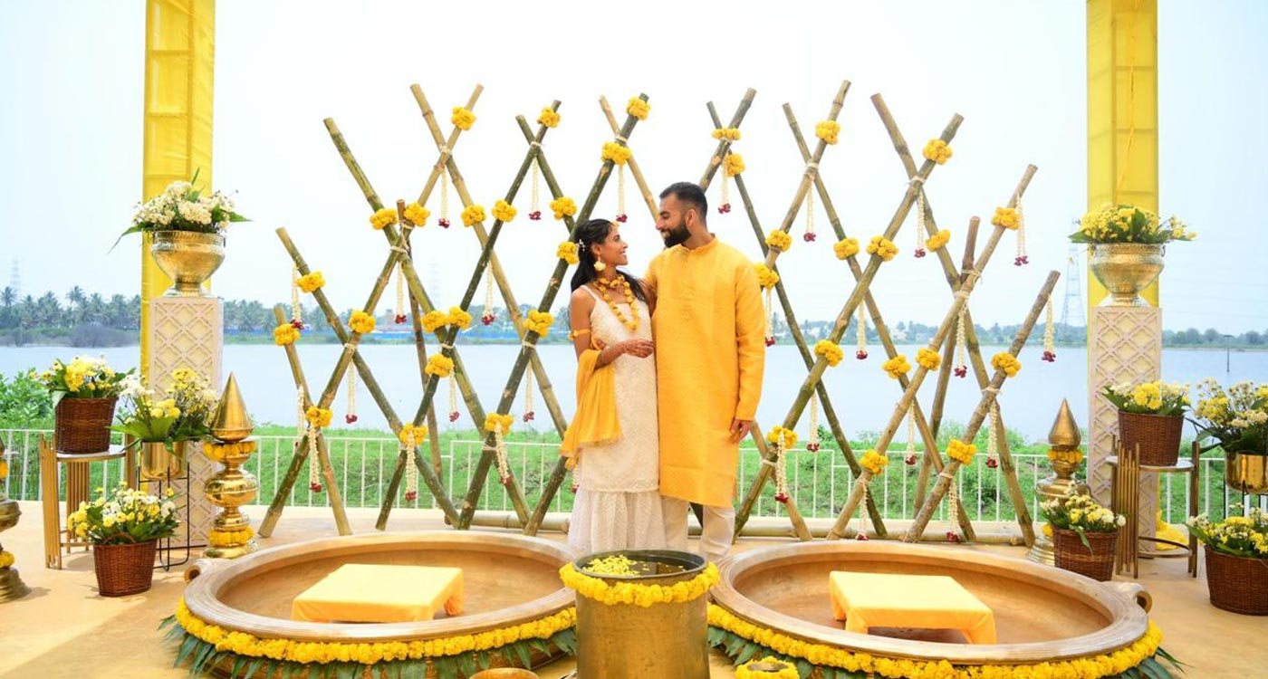 Haldi setup on Moongate's lakefront stage: a bamboo lattice backdrop hung with marigold and jasmine strings, two large brass urli bowls with cushioned seats in front, and the lake and far shoreline visible behind under flat mid-morning light