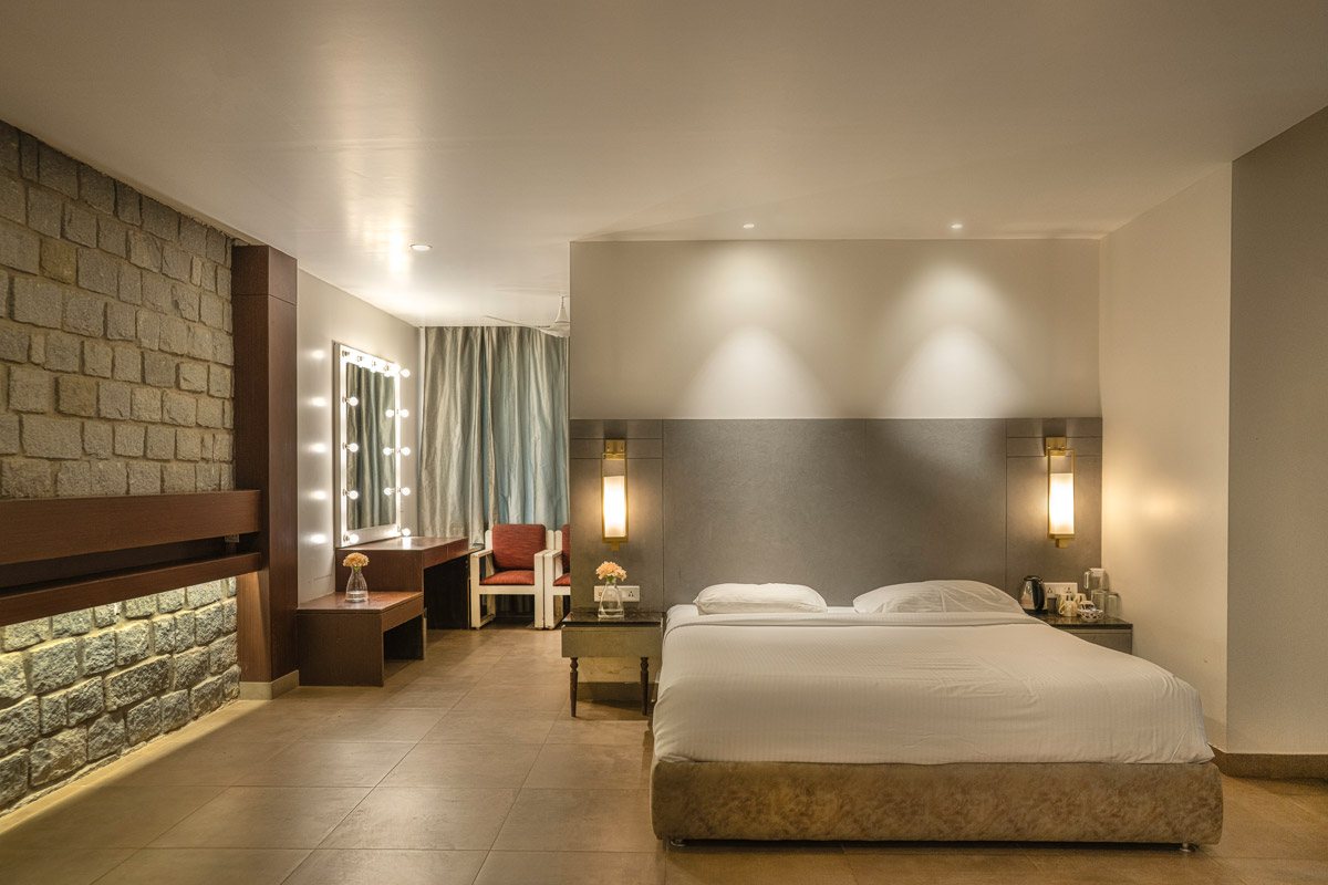 A Moongate suite interior — king bed against a grey upholstered headboard with brass wall lamps, an exposed stone-block feature wall down the left side, a bulb-lit vanity mirror and long desk, an armchair by the curtained window, and large-format stone floor tiles