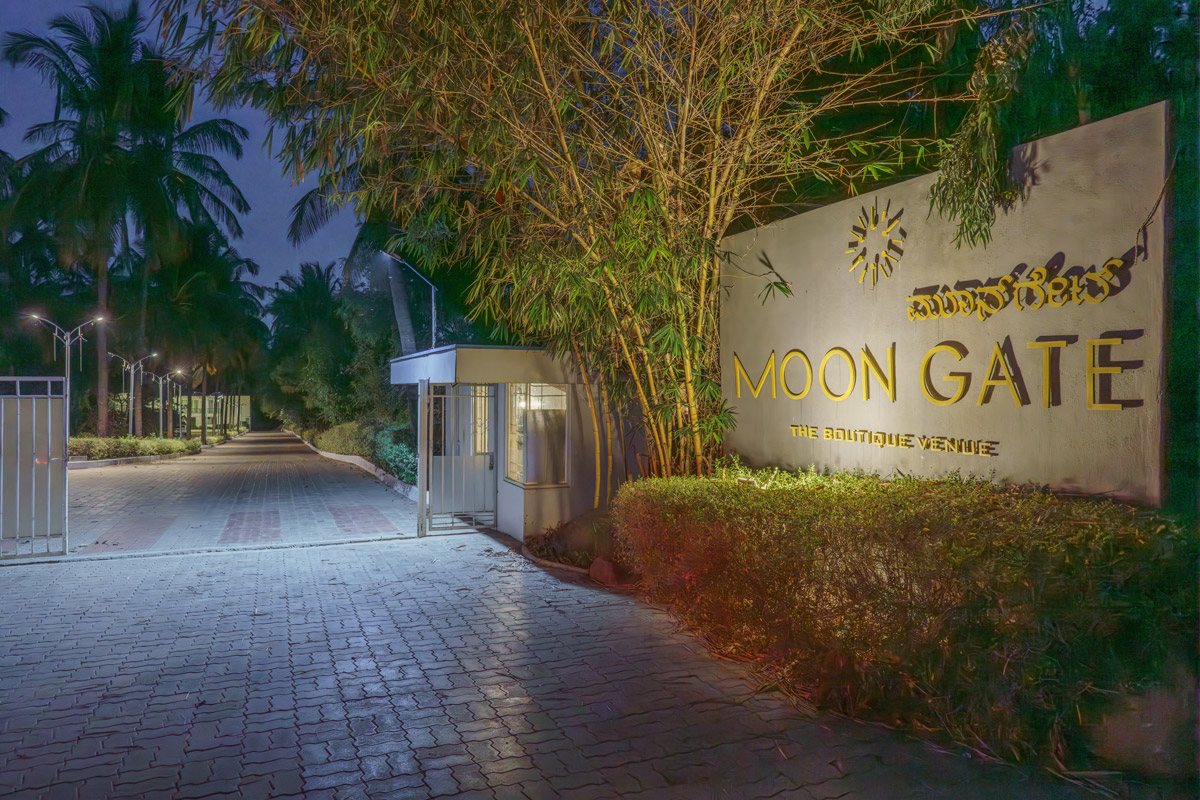 Moongate's entrance at dusk — the lit 'MOON GATE, The Boutique Venue' sign wall in gold lettering with the Kannada name above it, a security cabin and the paved driveway running away under street lamps between bamboo and coconut palms