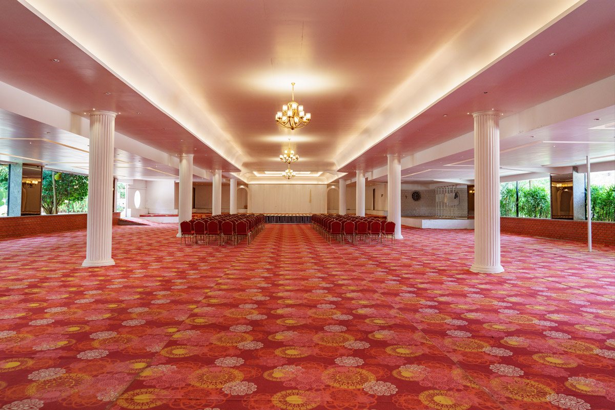 Opa Banquet Hall empty and set for theatre-style seating — red-and-gold patterned carpet running the full floor, a grid of fluted white columns, two brass chandeliers under a coved ceiling, and a block of red banquet chairs facing the stage wall at the far end