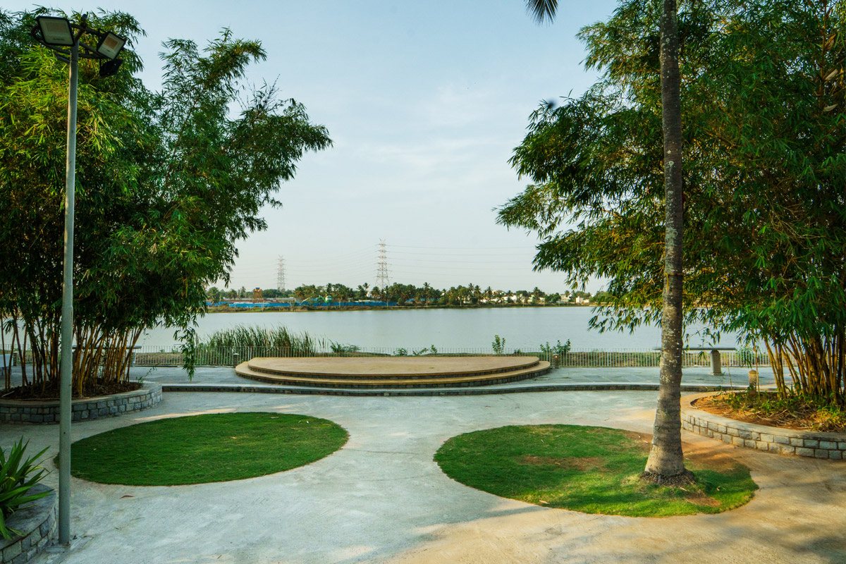 Lacu, the lakefront stage — a semi-circular paved platform of three broad steps set at the water's edge with the lake and far shore behind it, framed by bamboo clumps and a coconut palm, and two circular grass discs set into the paved apron in front, shot in afternoon light