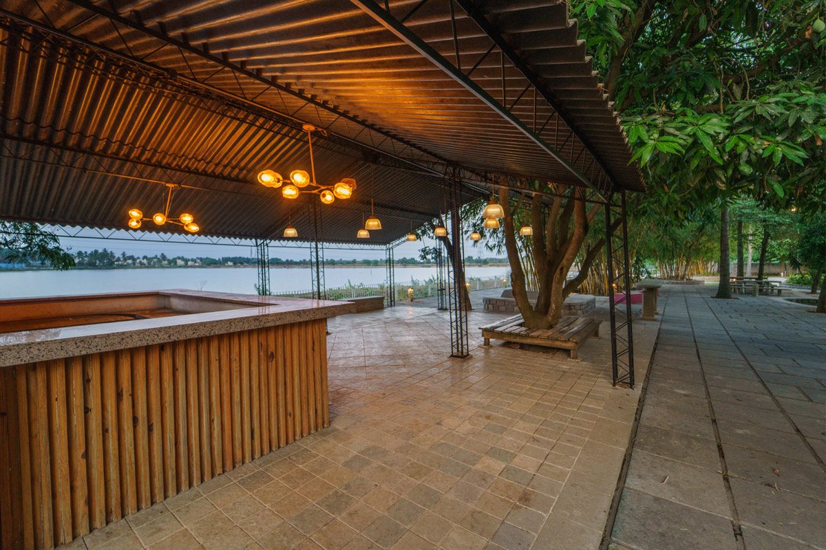 Covered lakefront deck at dusk — a long pitched metal roof on slim steel trusses over stone paving, a timber-clad bar counter in the foreground, brass globe pendants and a cluster chandelier lit warm, a mango tree growing through the deck with bench seating around it, and the lake beyond the open edge