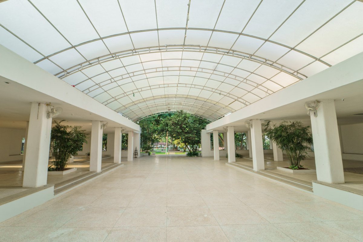 The covered courtyard walkway — a barrel-vaulted translucent roof on a steel frame throwing flat diffused daylight over a pale terrazzo floor, white square columns down both sides with planted bamboo between them, and the green lawn visible through the open end