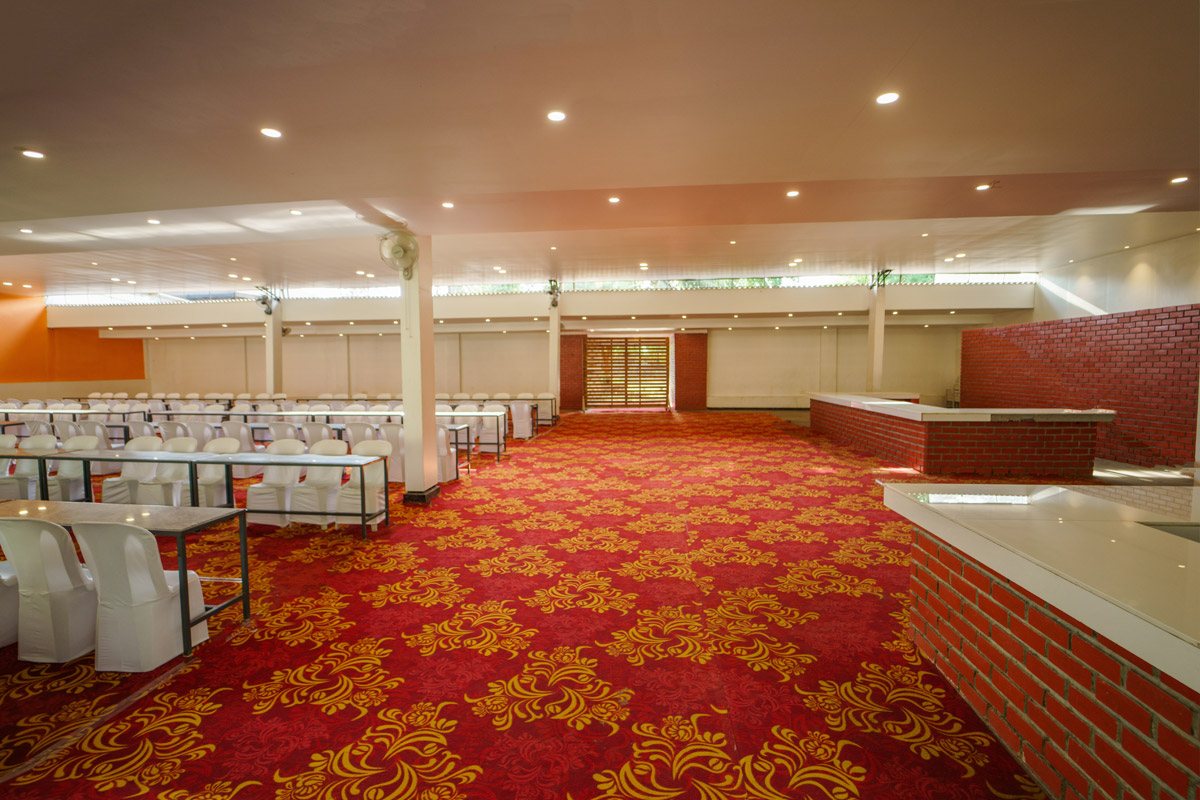The Courtyard Hall set for a seated function — red-and-gold patterned carpet, white banquet chairs in rows on the left, an orange accent wall, and permanent brick-faced buffet counters with white stone tops built along the right-hand side under recessed downlights