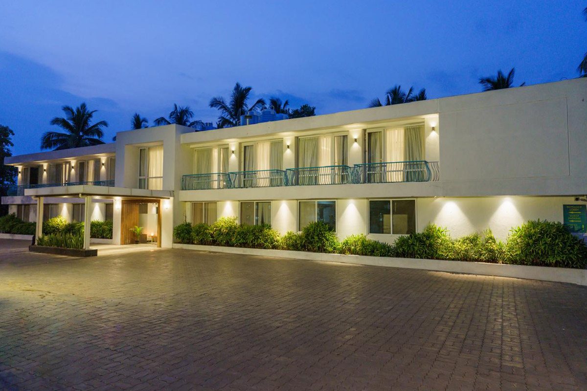 A two-storey suite block at blue hour — white rendered façade with warm wall-washer lighting under each window, teal metal balcony railings on the upper floor, a timber-lined entrance lobby lit from within, planting along the base and a wide paved forecourt in front