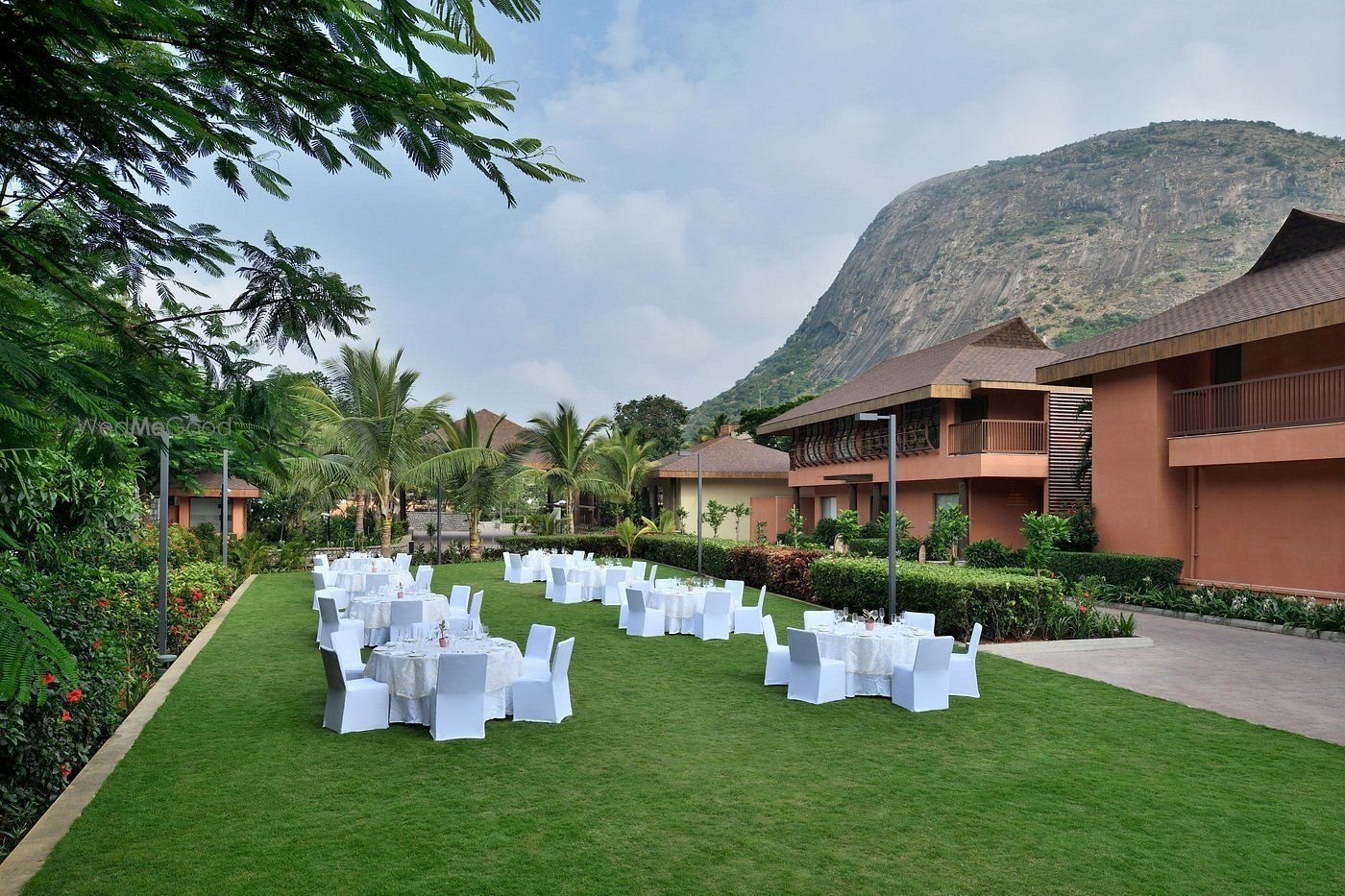 Shanti Lawn at Mulberry Shades set for a daytime meal — white-linen round tables with covered chairs on clipped grass, the terracotta guest-villa blocks along the right edge and the bare granite face of Nandi Hills rising directly behind under a partly clouded sky.