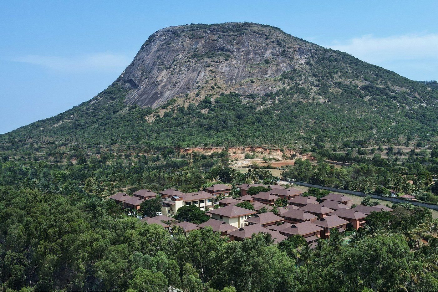 Aerial view of the whole resort in midday light — clusters of shingled pyramid roofs over terracotta bungalows set in dense green, with the rounded granite dome of Nandi Hills filling the frame immediately behind the property.