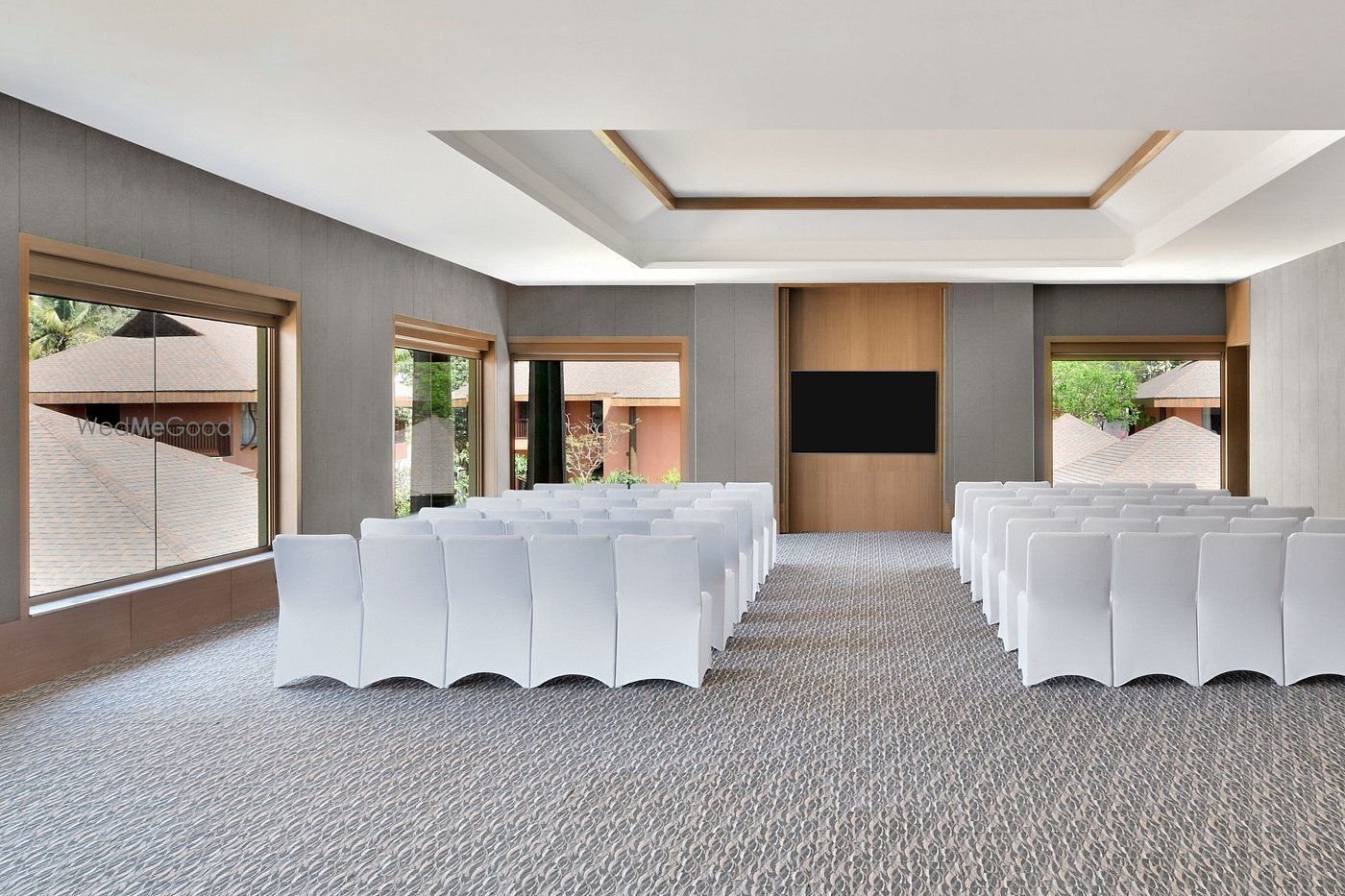 The Amaya indoor hall set theatre-style in daylight — rows of white-covered chairs on patterned grey carpet, grey fabric walls, a shallow recessed ceiling, a timber AV panel with a mounted screen on the end wall, and tall picture windows on both side walls looking onto villa roofs and trees.