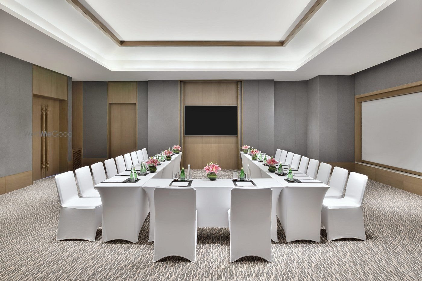 The inner half of the Amaya hall in a U-shape conference set — white-skirted tables with pink lily centrepieces, the same patterned grey carpet and recessed ceiling, a timber AV panel with mounted screen on the far wall, a pull-down projection screen on the right and a gold-handled double door on the left; no windows in this half.
