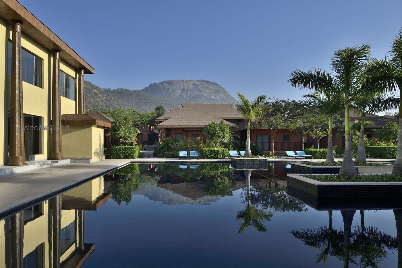 The long reflecting pool in early-morning light, mirroring a two-storey ochre block with tall columns on the left, a row of single-storey villas along the far edge, royal palms on the right and the Nandi Hills ridge behind under clear blue sky.