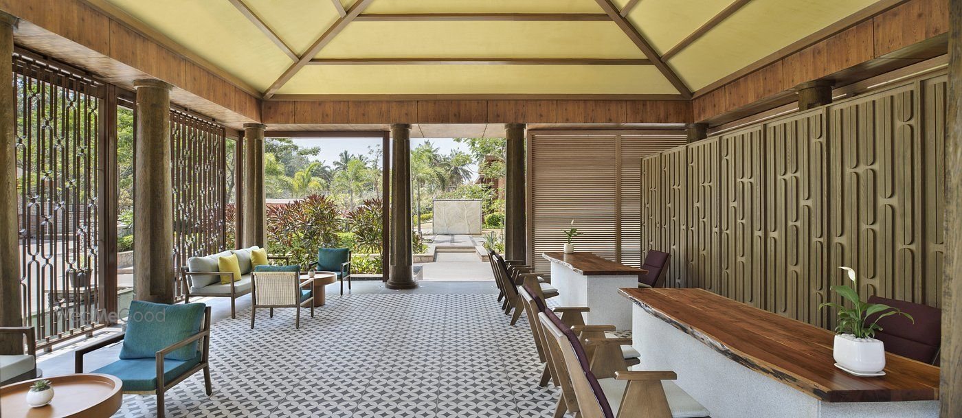 An open-sided pavilion lounge — live-edge timber bar counters with high-backed stools on the right, low lounge chairs and a sofa on the left, carved timber screens along the walls and a translucent yellow pitched roof over a black-and-white patterned cement-tile floor, open on two sides to the garden.