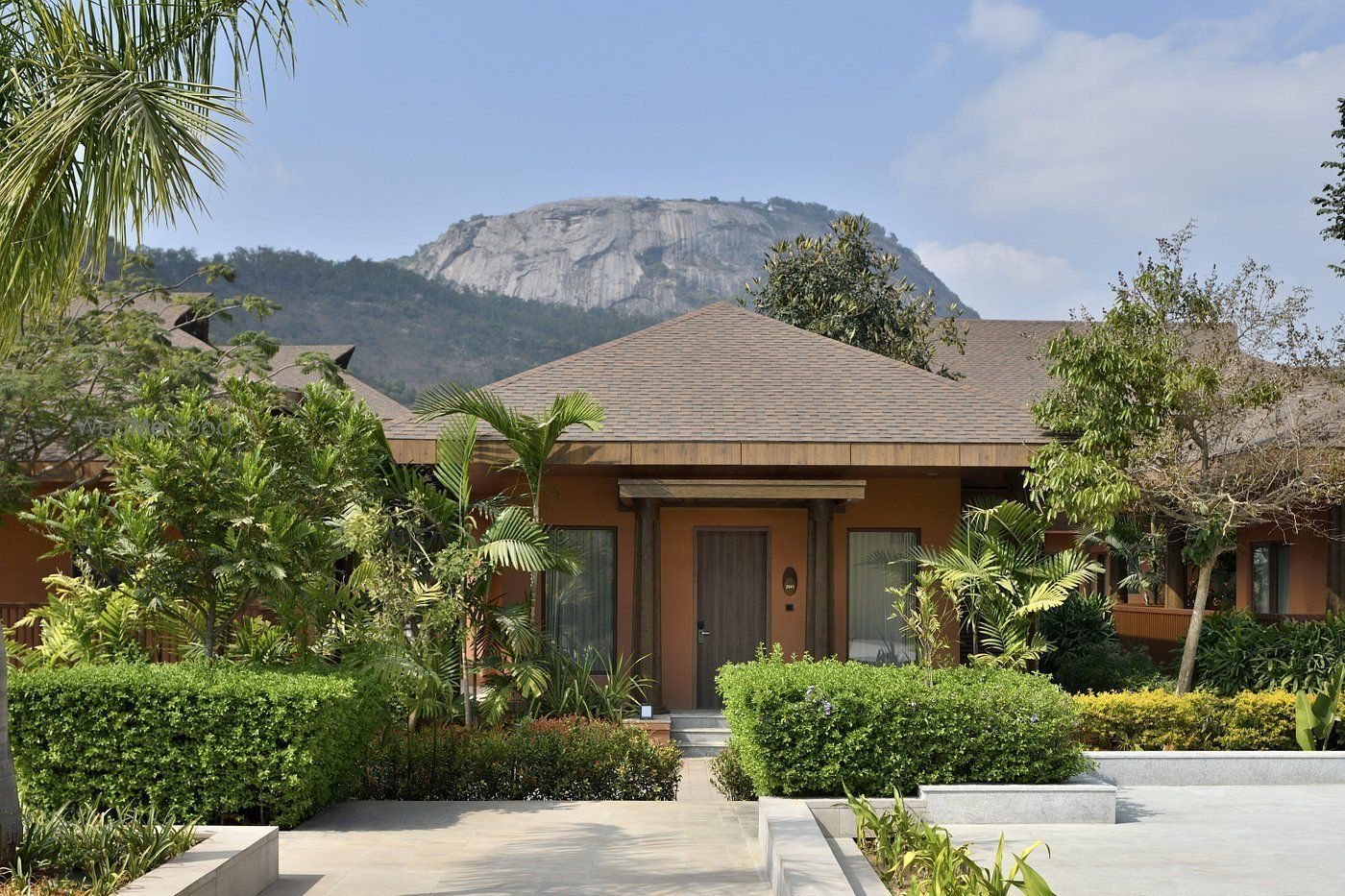 Front elevation of a single-storey guest bungalow in daylight — terracotta-washed walls, a shingled pyramid roof with a deep timber eave and a timber entrance door, framed by clipped hedges, palms and flowering beds, with the granite face of Nandi Hills rising behind the roofline.