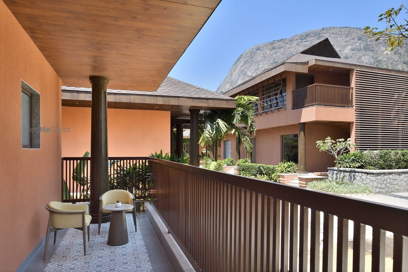 A first-floor open corridor of a guest block — two cane-backed armchairs and a small round table on patterned grey tile under a timber soffit, a slatted timber railing, looking across a planted courtyard to a two-storey ochre villa block with the Nandi Hills rock face behind it in midday sun.