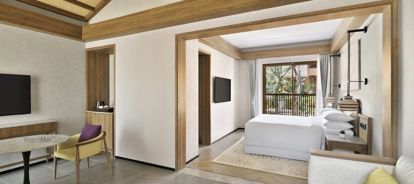Interior of a bungalow guest room — a white-linen king bed under a timber-framed archway with wall-mounted reading lights, a writing desk and armchair in the foreground, pale herringbone flooring, and a glazed door opening onto a balcony with greenery beyond.
