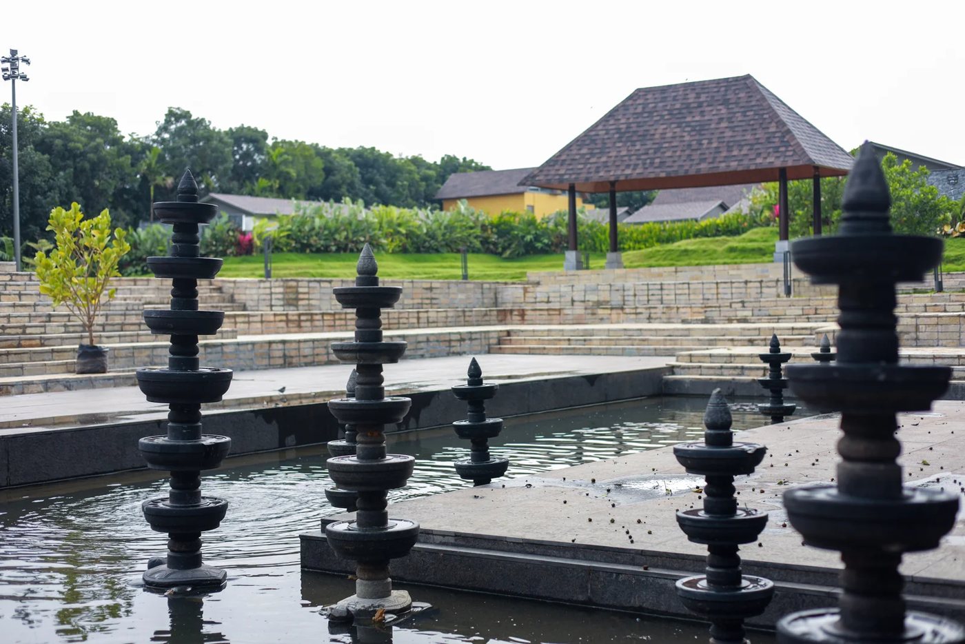 The Kalyani water tank at Nature Knots — tiered black stone lamp columns standing in the water, with the stone terrace stepping up behind it to a tiled-roof open pavilion and lawn