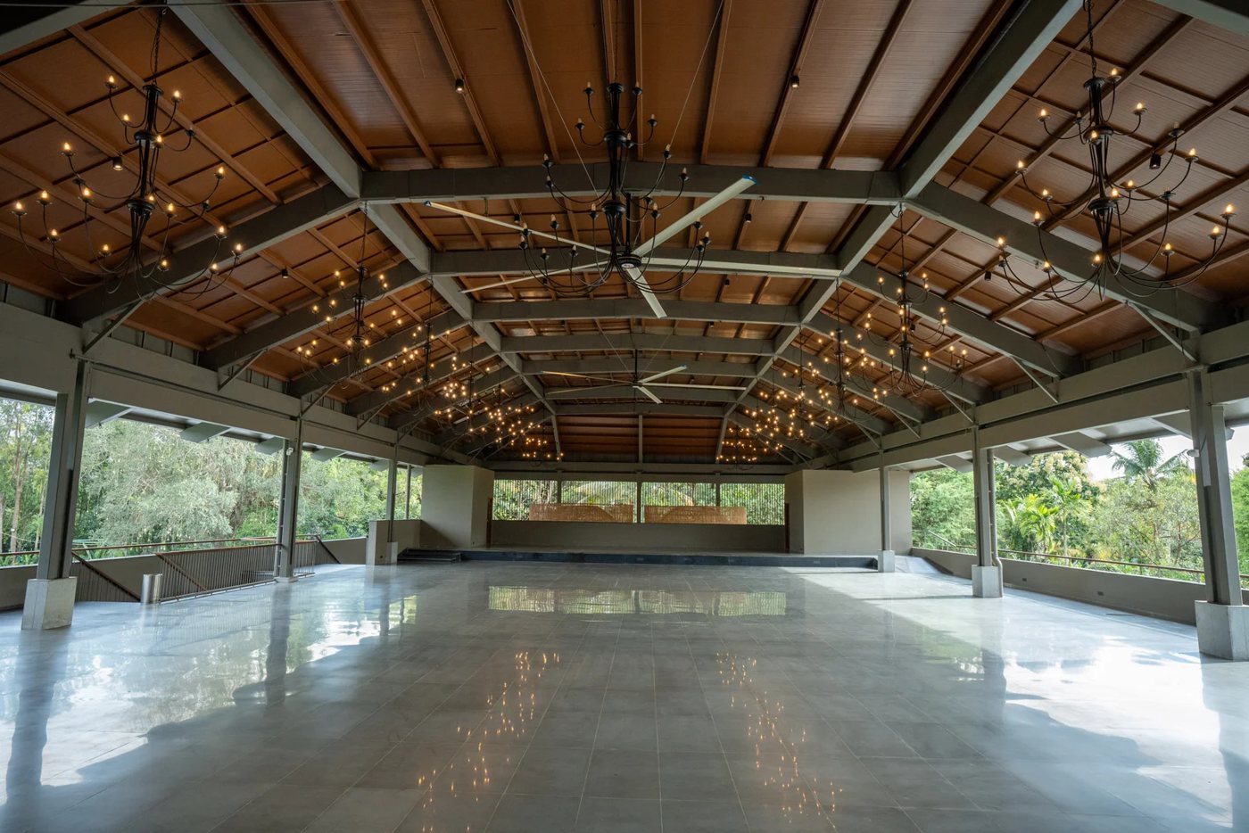 Kedar, the covered banquet hall at Nature Knots — a pitched timber-lined roof on steel trusses, open sides looking onto greenery, festoon lights and ceiling fans over an empty polished floor
