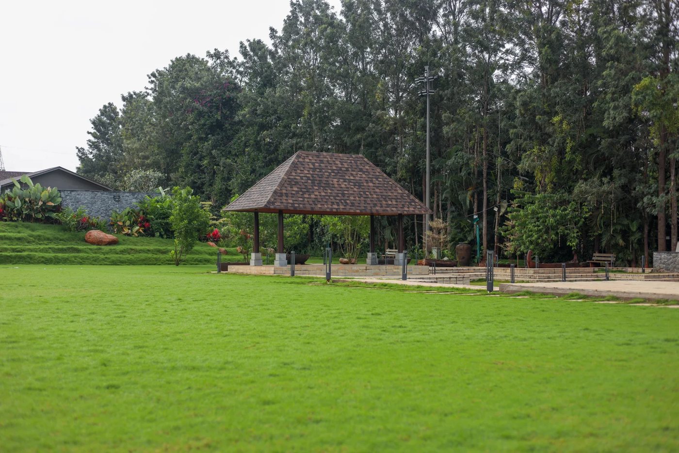 A wide level lawn at Nature Knots with a tiled-roof open pavilion at its edge and a line of tall mature trees behind