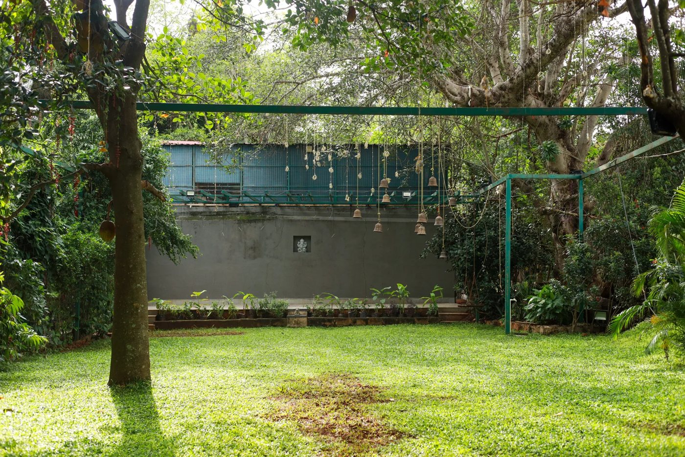 Abheri at Nature Knots — a small enclosed lawn beside a grey wall with a mature tree and a row of hanging terracotta bells strung on a frame overhead