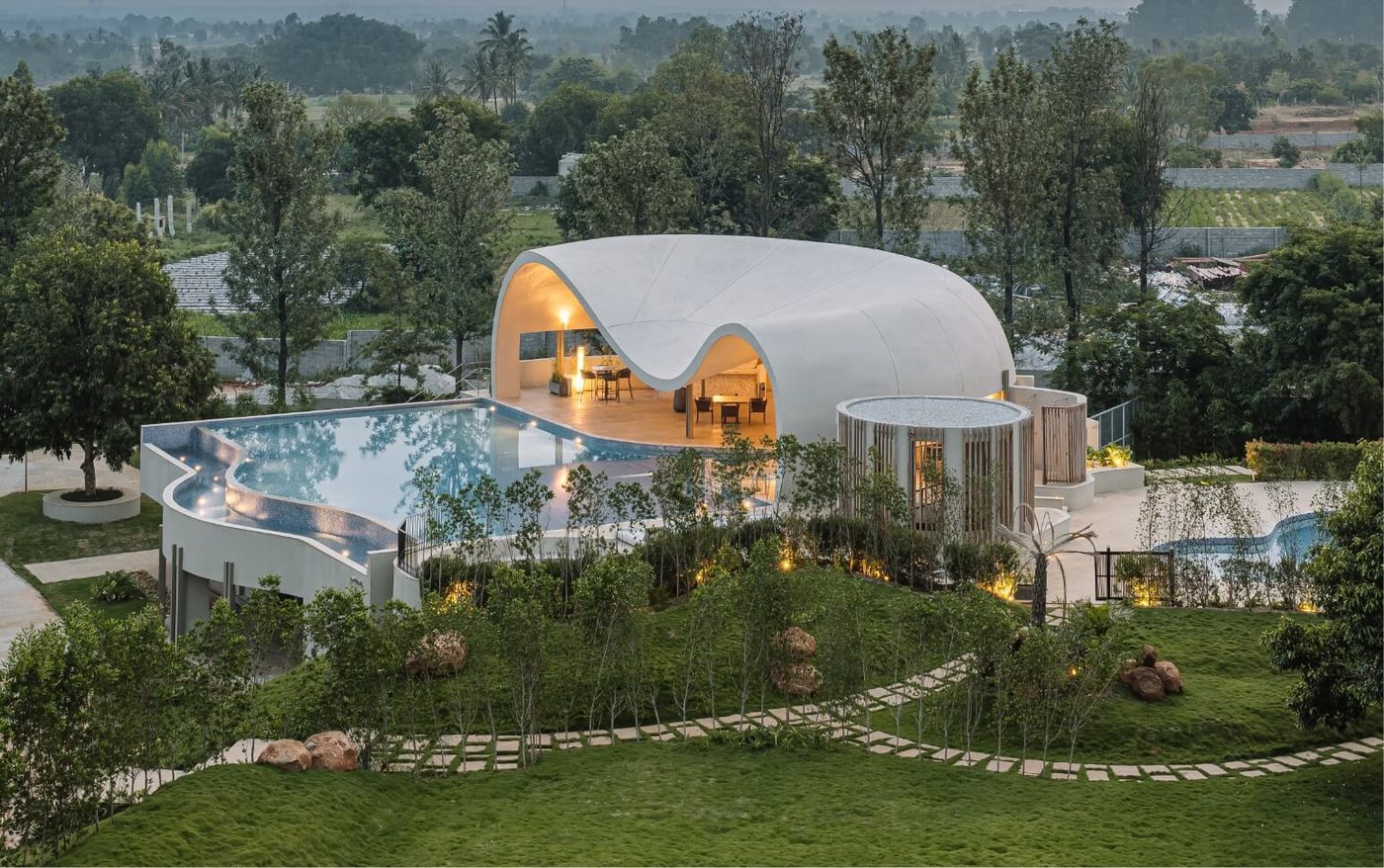 ORO The Estates at dusk from above — the free-form white concrete shell of the Aqua clubhouse pavilion curling over a lit lounge, the infinity pool wrapping around it, a circular timber-battened jacuzzi rotunda alongside, and the landscaped lawn below threaded with stepping-stone paths and buried lighting, with the Nandi Hills farmland stretching to the horizon.