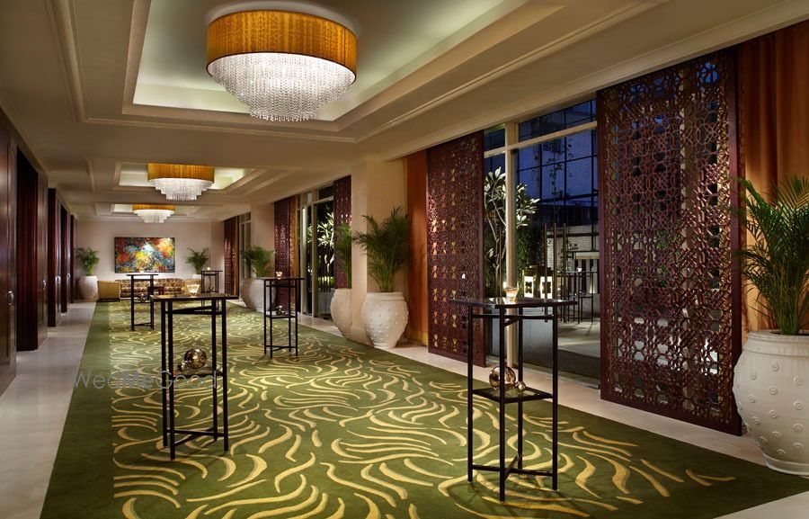 The ballroom pre-function corridor at The Ritz-Carlton Bangalore — green patterned carpet, drum chandeliers, carved dark jaali screens between full-height windows and white planted urns